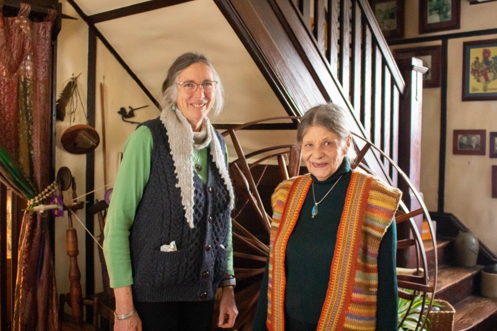 Two older white women stand together smiling and wearing hand made clothing.