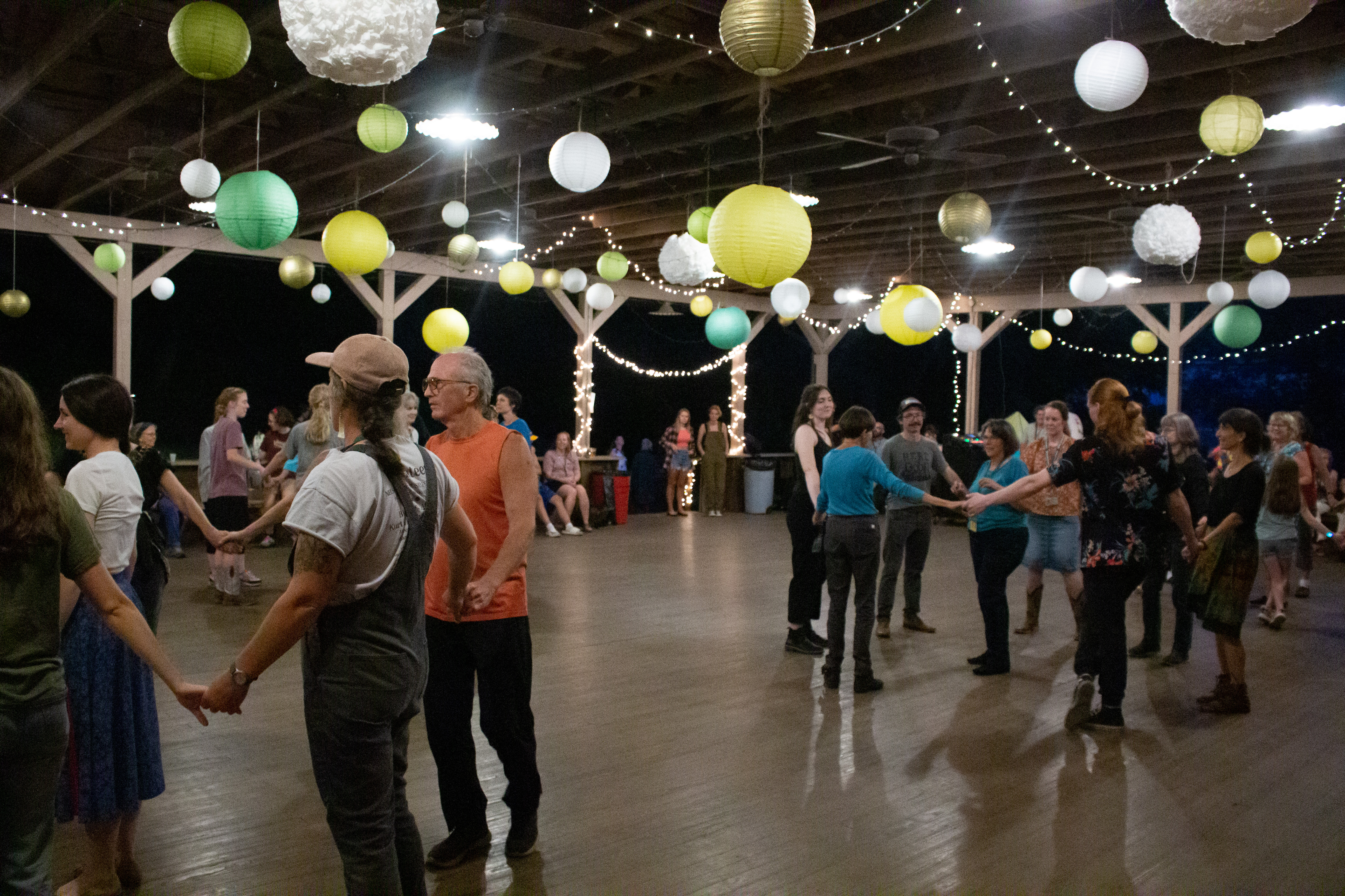 An open air dance pavillion with wooden floors and colorful paper ball ceiling ornaments and twinkle lights. Dancers hold hands in a circle, multiple circles of dancers.