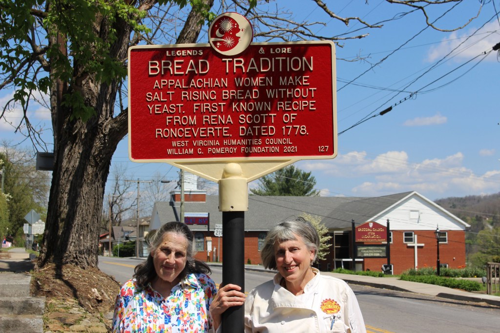 A red marker on a street above two women. The marker reads "Bread Tradition" with body text that says "Appalachian Women make salt rising bread without yeast. First known recipe from Rena Scott of Ronceverte. Dated 1778."