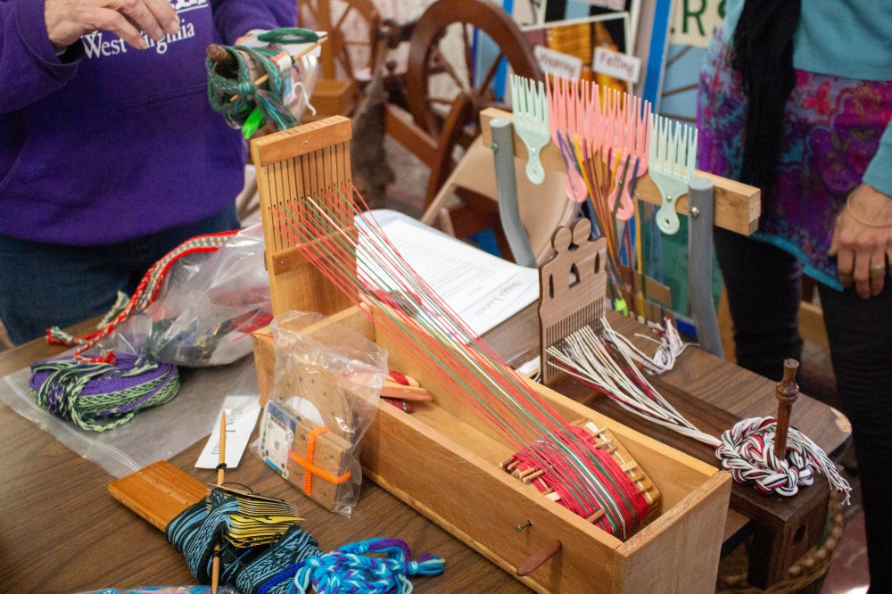 Several varieties of small looms, some made from wood, some from found materials, sit on a table and each hold works in progress.