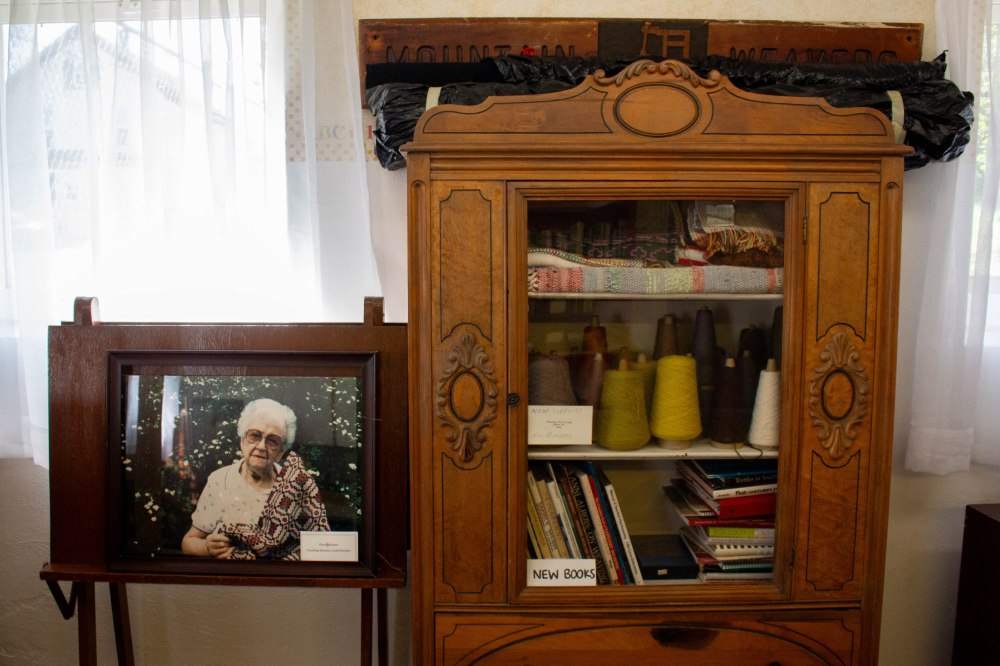 A framed photograph of an old woman with a woven item sits to the left of a large wooden cabinet of books, yarn, and fiber products.