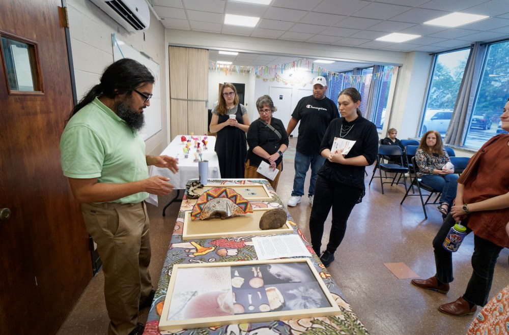 A man with dark hair and a green shirt looks down at a table with mushroom-related information and artist conks as he speaks to a gathering crowd around his table.