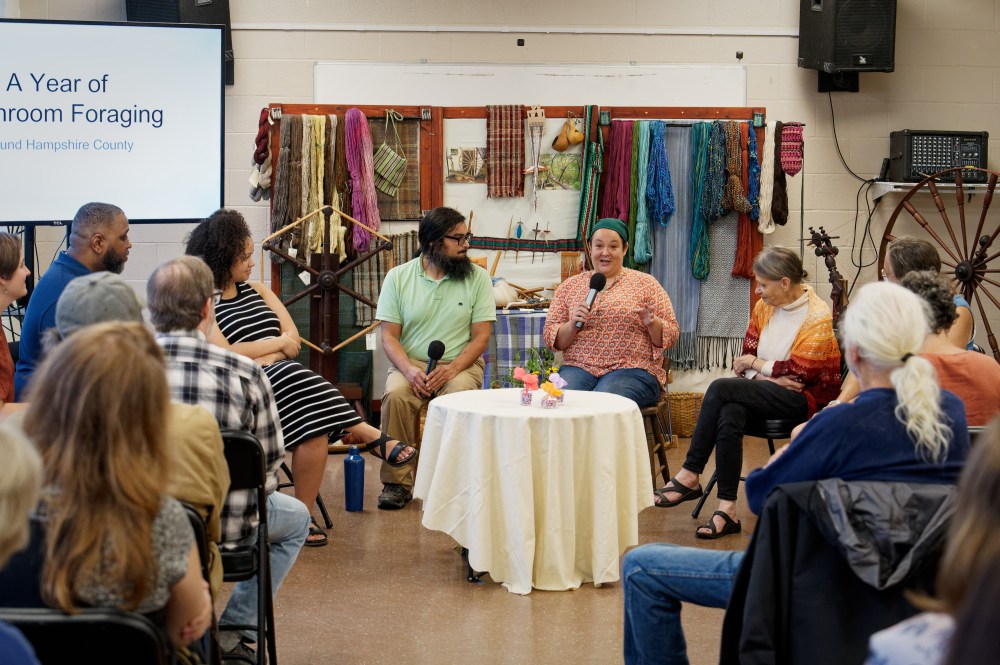A man and woman hold microphones and speak to an audience with co-presenters sitting beside them. In the background, a screen reads "A Year of Mushroom Foraging" and a display of fiber arts and yarns. In the foreground, the audience watches.