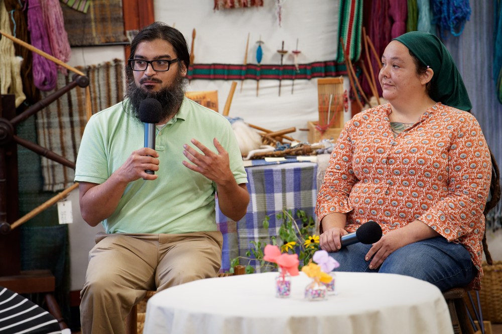 A man with dark hair and glasses and a green shirt holds a microphone and speaks to an audience with his mentor beside him, a woman wearing a green hair scarf and orange patterned shirt, also holding a microphone and listening.