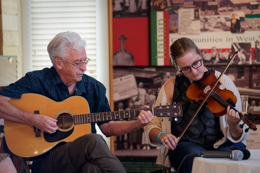 An older man plays acoustic guitar with a younger woman playing fiddle.
