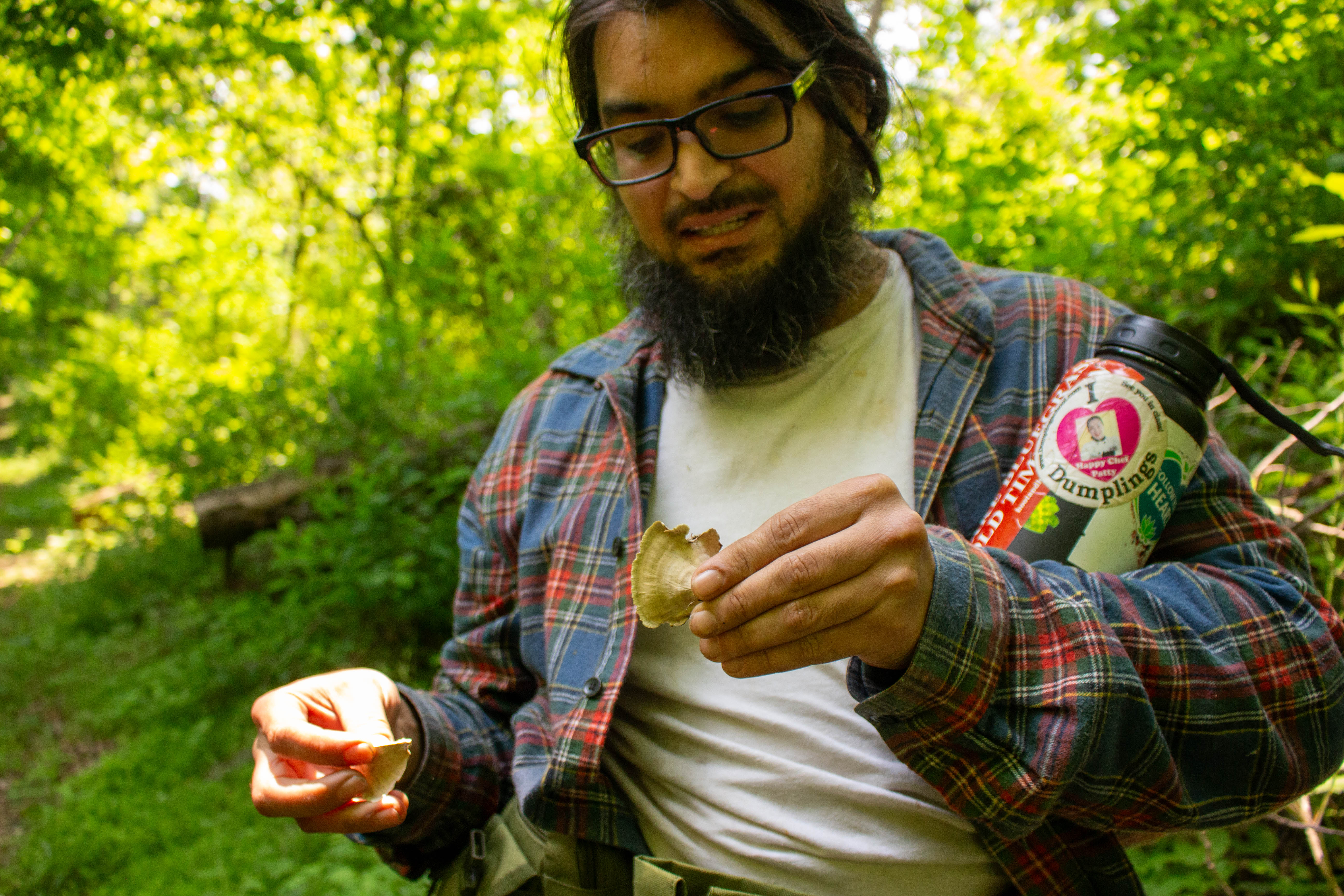 A man holds his water bottle in his arm and holds small mushrooms in his hands while outdoors.