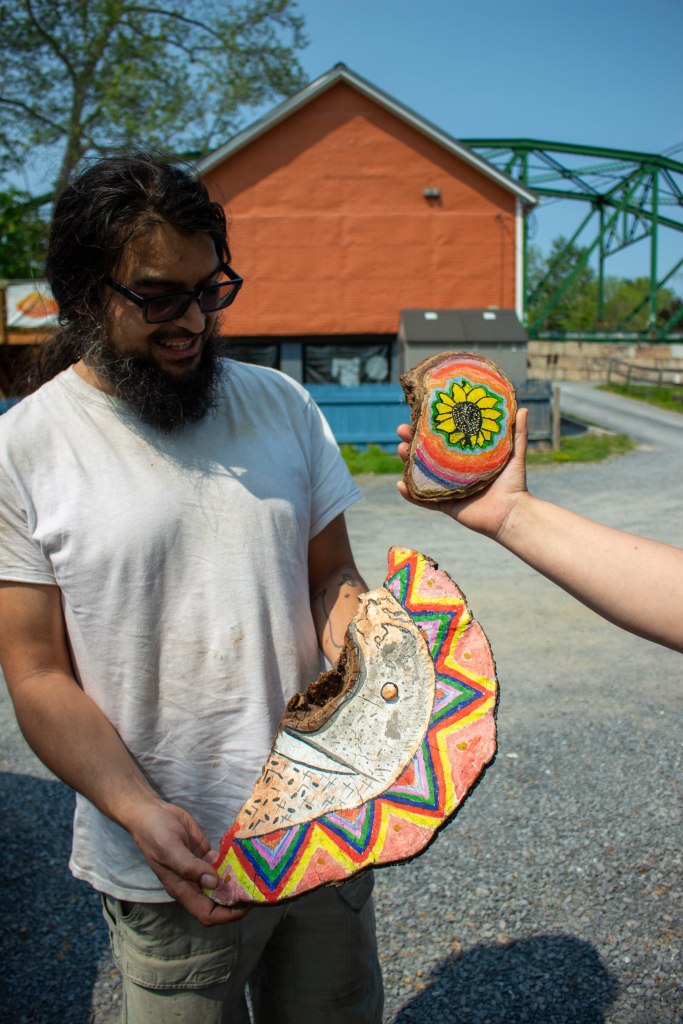 A man with glasses and dark long hair and a beard with a white t-shirt holds a large colorfully painted mushroom "artist conk" while a hand holds out another painted example.