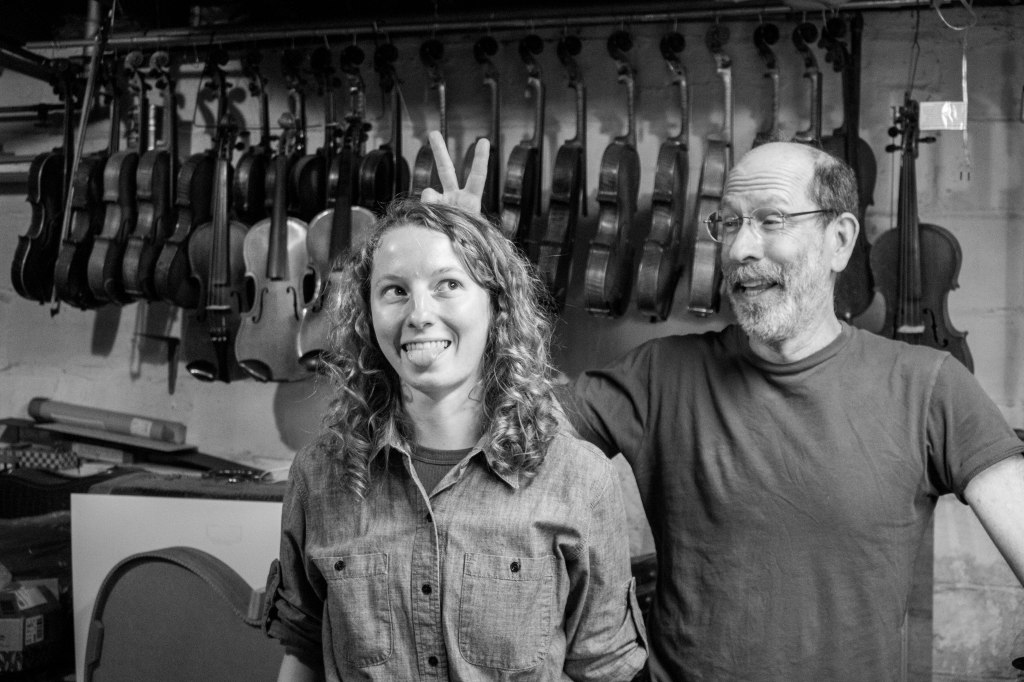 A young woman and older man in a basement with fiddles hanging along the wall. The man is making bunny ears over the woman's head as she sticks her tongue out for a goofy picture.