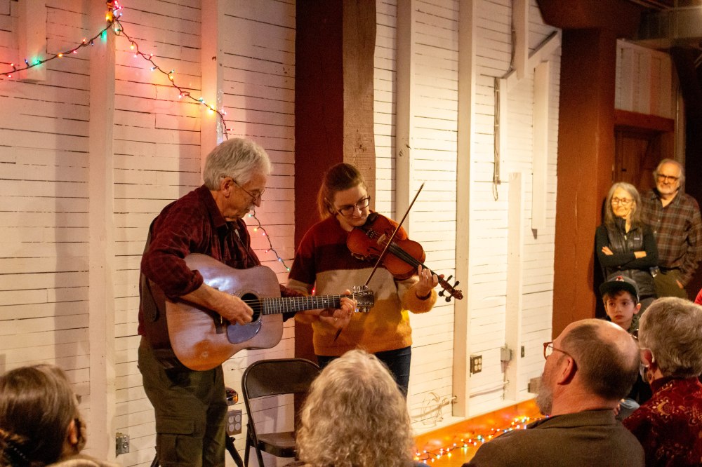 An older man plays guitar with a younger woman playing fiddle at an event for an audience.