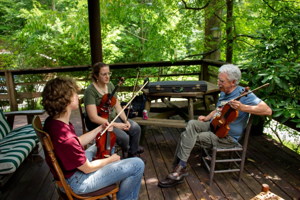 Two women sit with an older man outdoors on a porch, all holding fiddles.