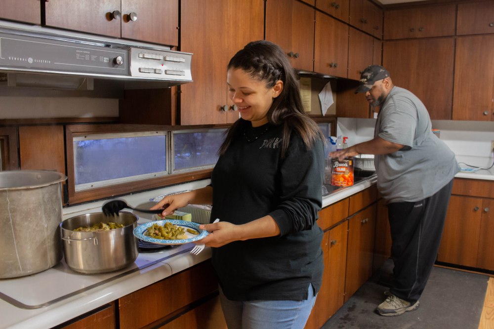 A Black woman with long hair and wearing a black long sleeved shirt smiles and scoops green beans onto her plate.