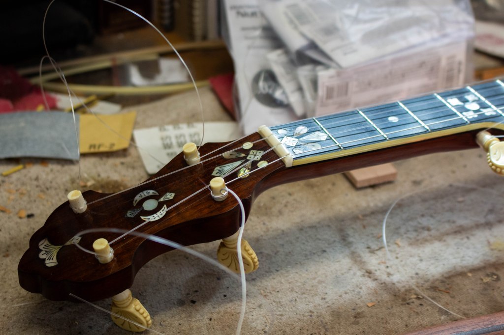 A vintage banjo with detailed inlays laid on a workbench, recently restrung and the strings are not yet trimmed on the pegs.