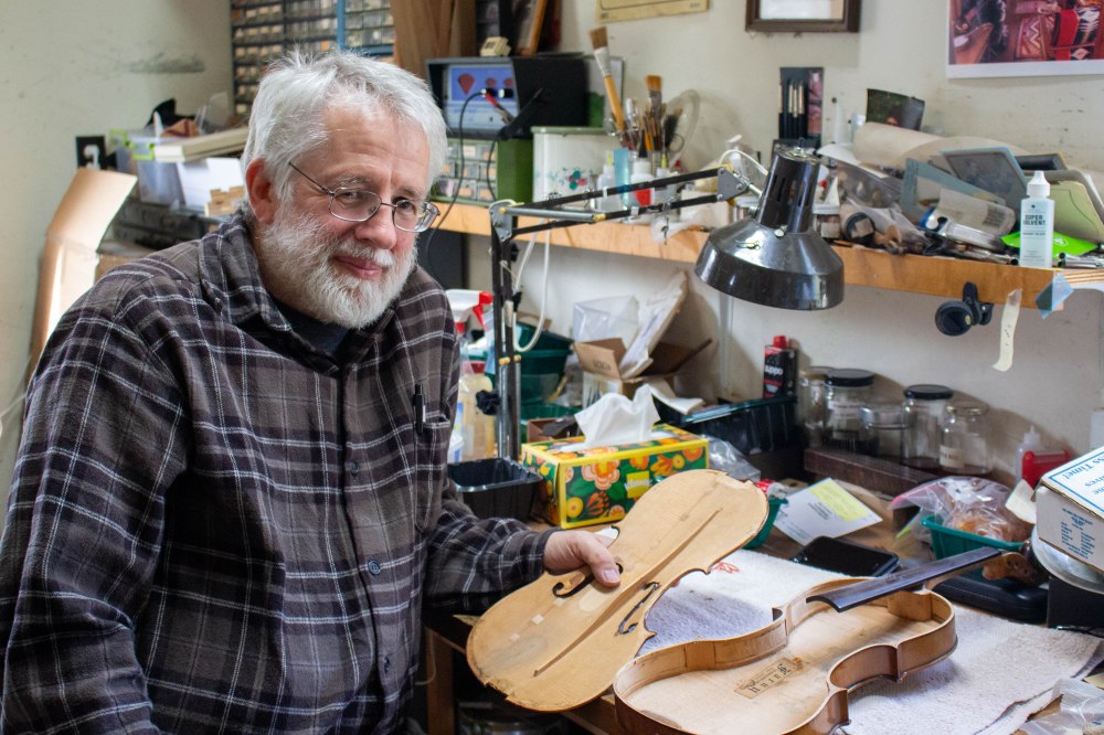 An older white man wearing a gray plaid shirt holds a piece of a violin in the process of being repaired, while sitting at his workbench.