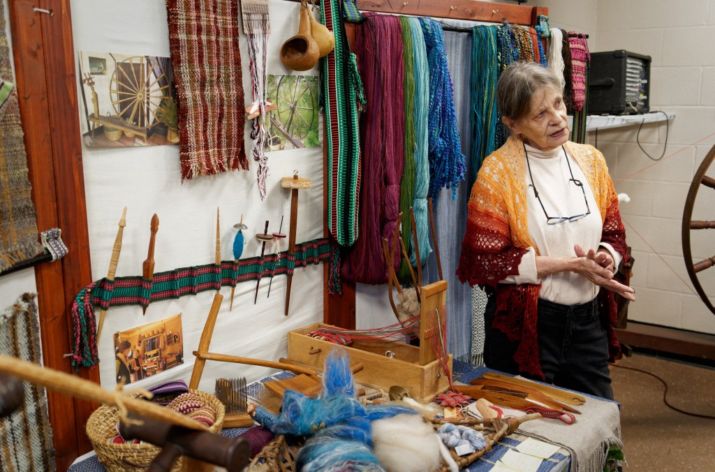 An older white woman stands beside a display of fiber arts tools and materials.