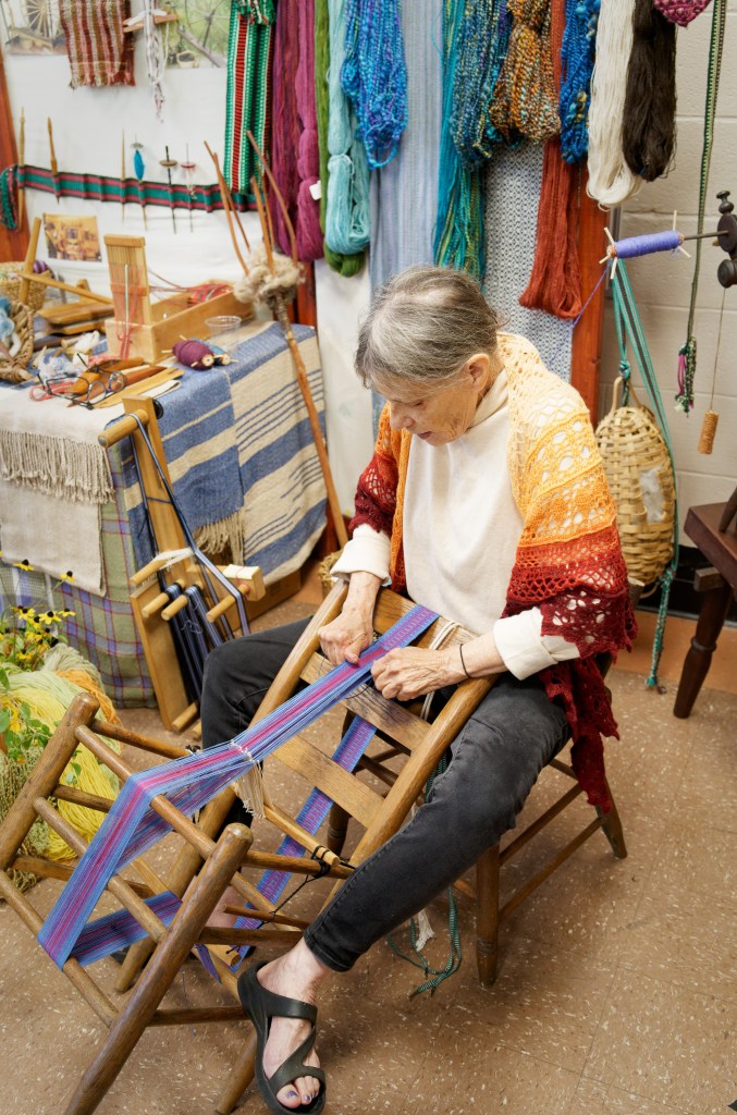 An older woman sits while holding a ladder back chair against her lap. The ladderback chair is warped as a loom with blue and purple thread. The woman is demonstrating how to weave a band using this traditional method.