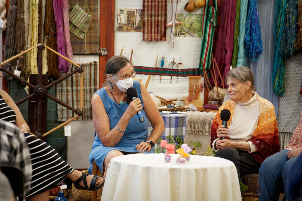 Two older white women sit together with microphones, a fiber arts display behind them.