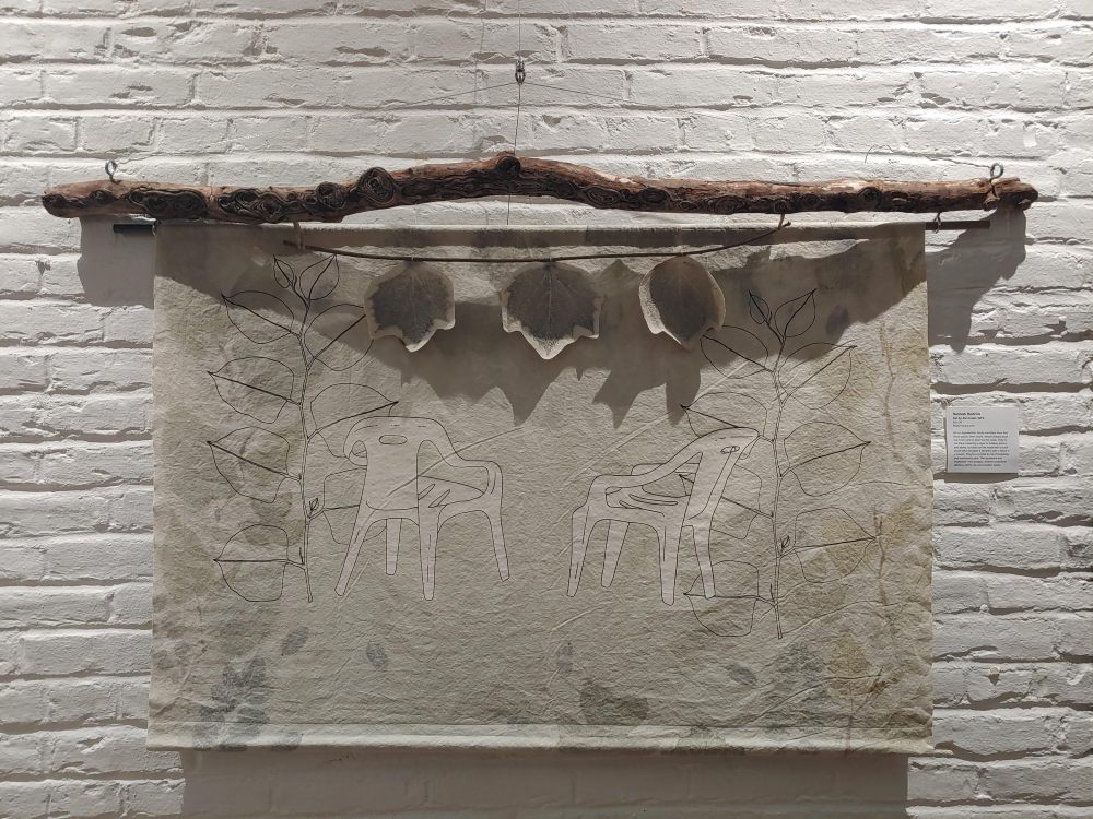 An art piece hanging against a white painted brick wall. A found wooden stick holds up a rectangular cloth with eco-printed designs and two screen printed white plastic chairs in the center.