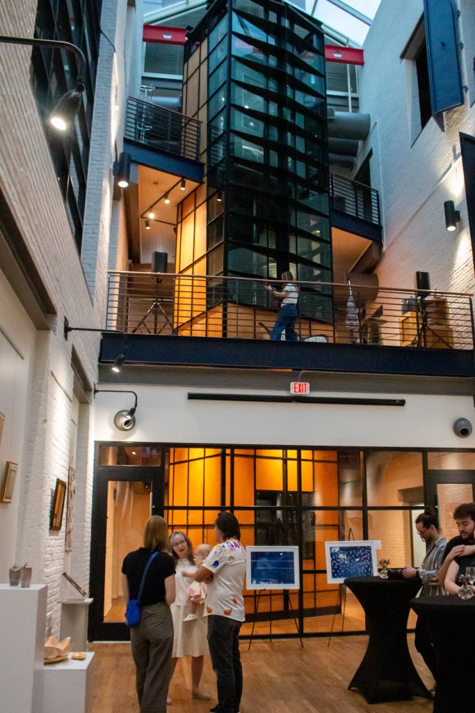 A view of the exhibit space located in the Wheeling Artisan Center. An open room with several windows, white painted brick, art works displayed on the ground floor, and a view of the second and third floors above.