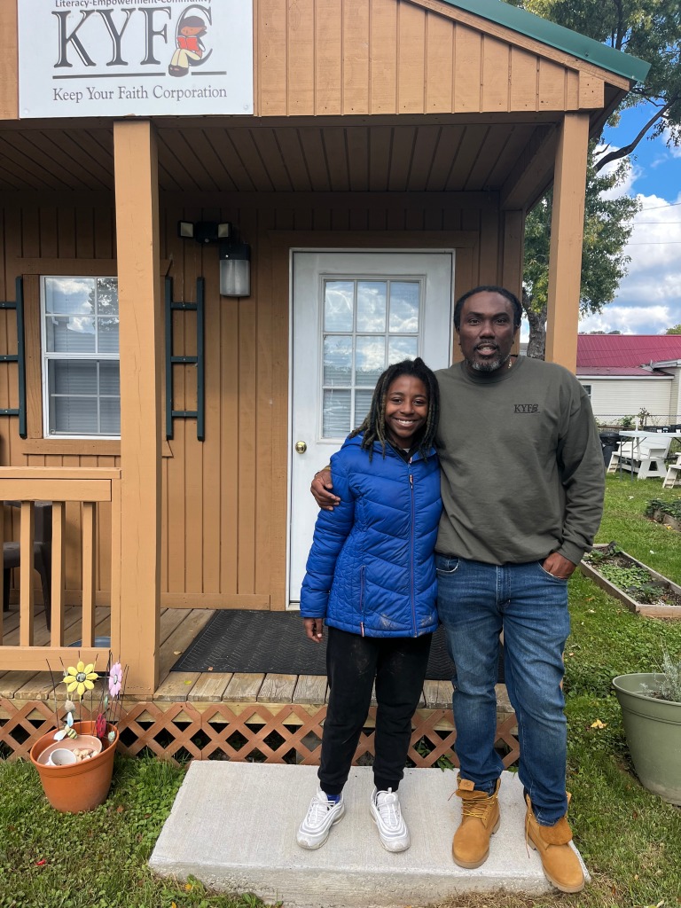 An older Black man in a gray long-sleeved shirt stands with his arm around a younger Black woman wearing a bright blue puffy jacket, both smiling for the camera. They are standing outside in front of the Keep Your Faith Corporation building and their urban farming plot.