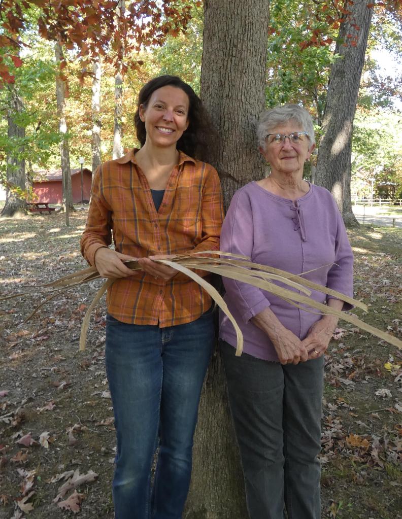 Two white women stand against a tree outside smiling together. On the left, a younger woman with brown hair wearing an orange flannel button down and holidng long splints from white oak used to make baskets. On the right, an older woman with white hair and glasses wearing a purple sweater.