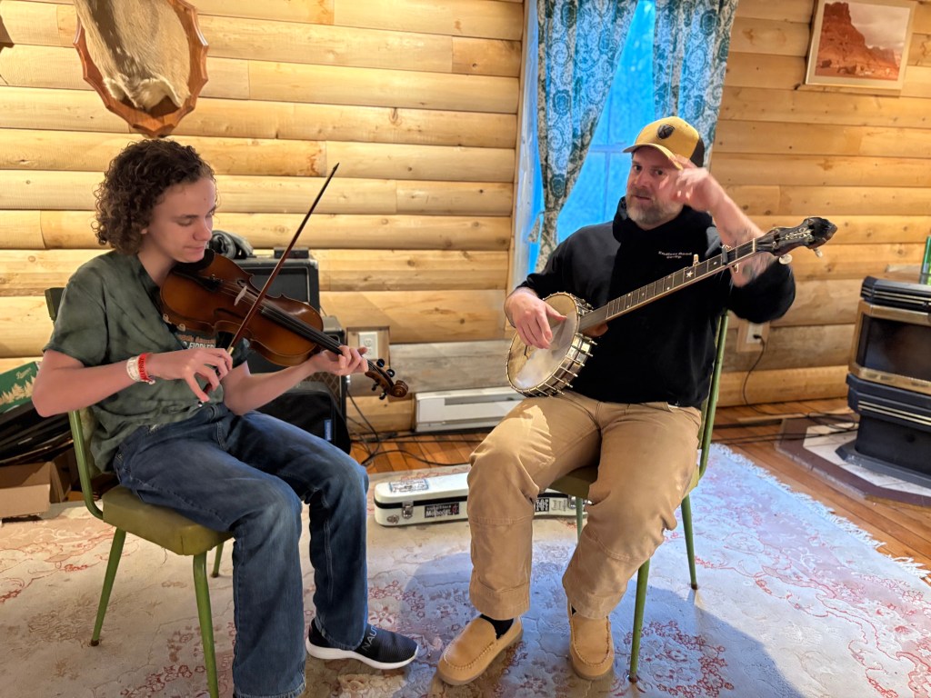A teenage boy playing fiddle and sitting beside a man playing the banjo.
