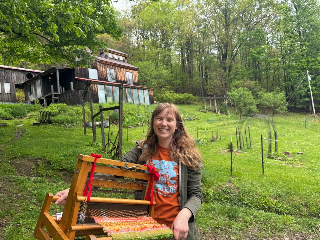 A woman wearing a red shirt and sweatshirt is holding a loom and smiling, standing outside a homestead on a hill.