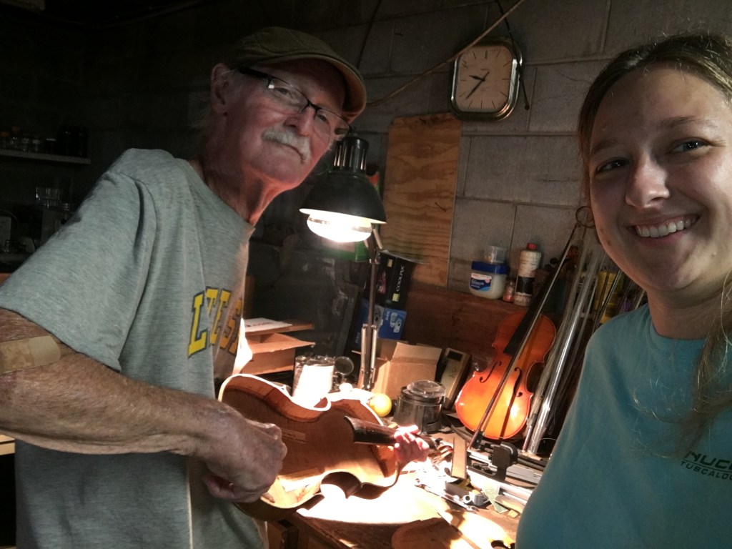A man and a woman pose for a selfie beside a work bench with a fiddle and tools.