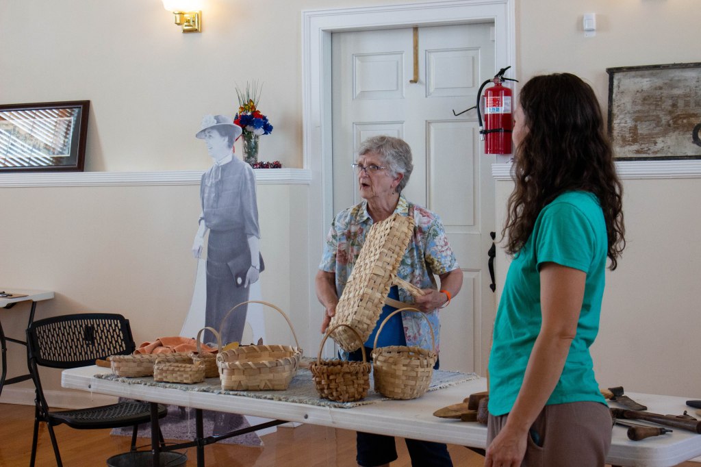 Two women stand beside a table filled with baskets as the older woman holds up a basket and addresses the audience.