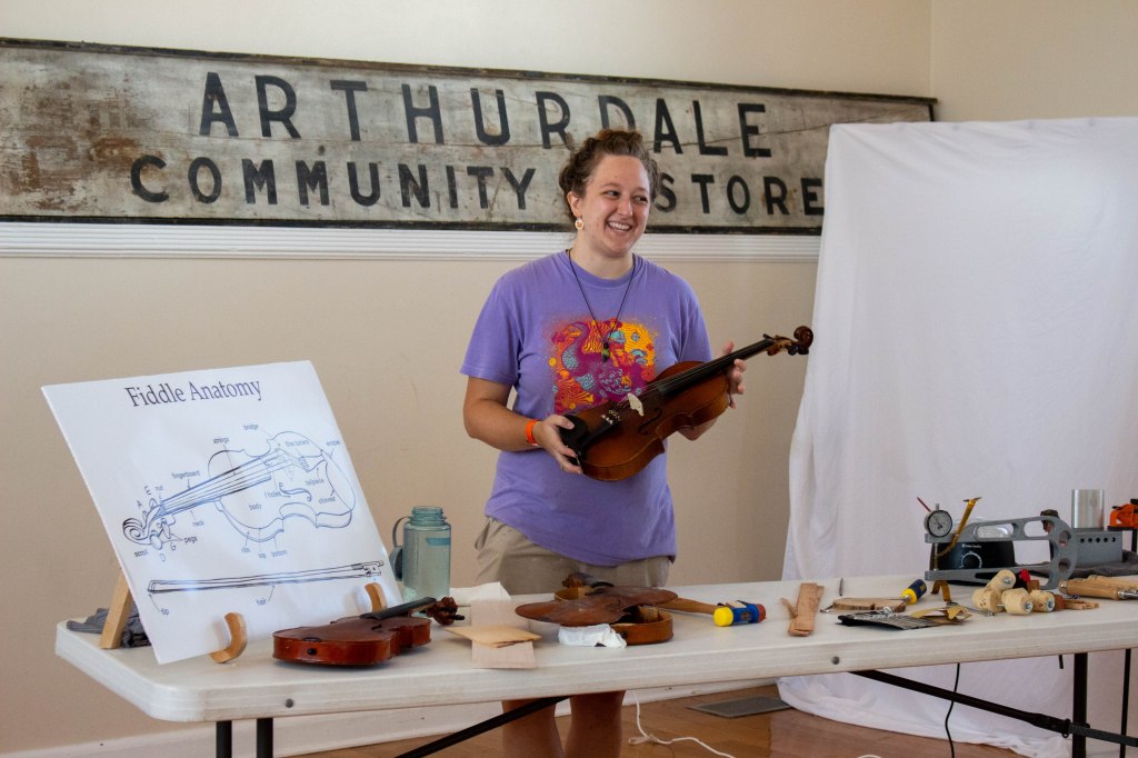 A woman in a purple t-shirt holds a fiddle and stands beside a table filled with broken fiddle pieces and tools.