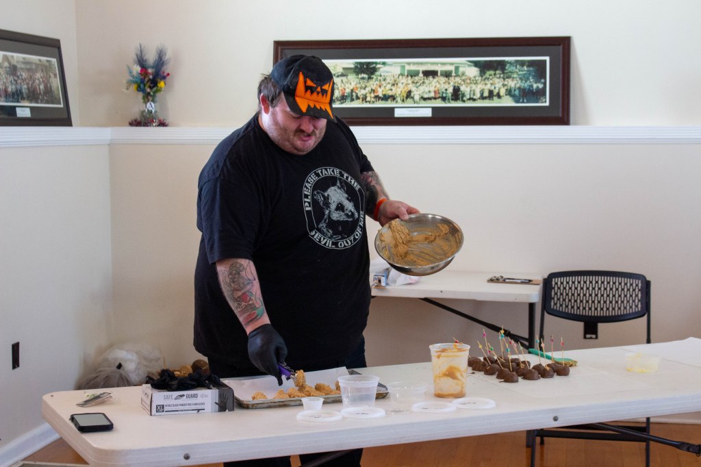 A man in a black t-shirt and hat stands beside a table and demonstrates making chocolate dipped peanut butter candies.