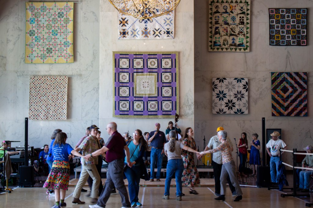 A crowd of people square dancing. The wall behind them is lined with beautiful quilts. The band plays in the back ground.