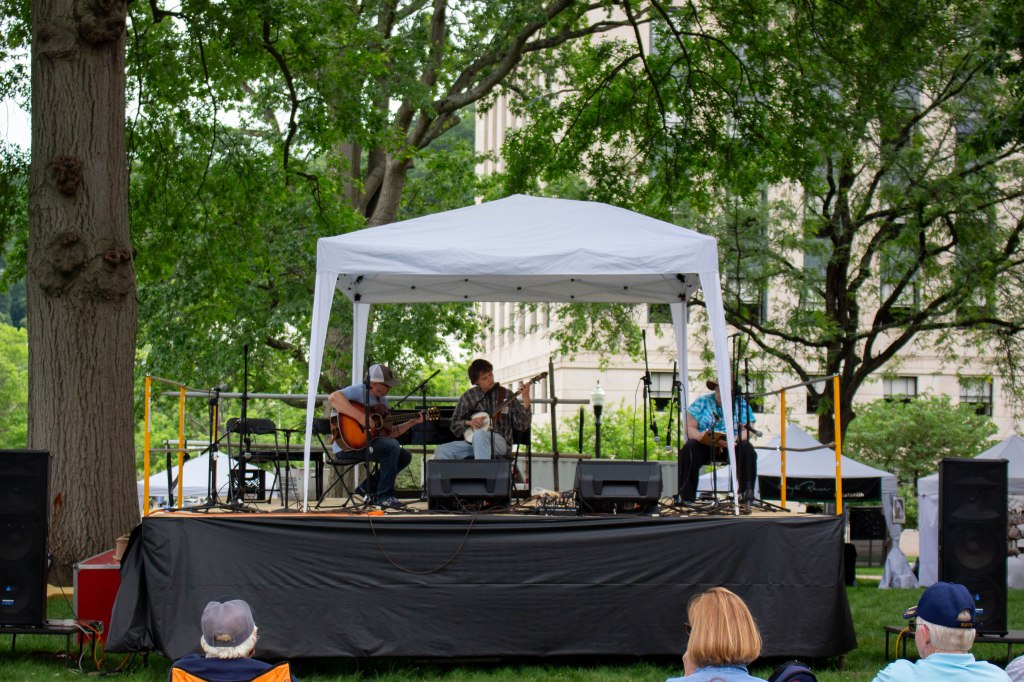 Two musicians play on an outdoor stage with a white tent overhead.