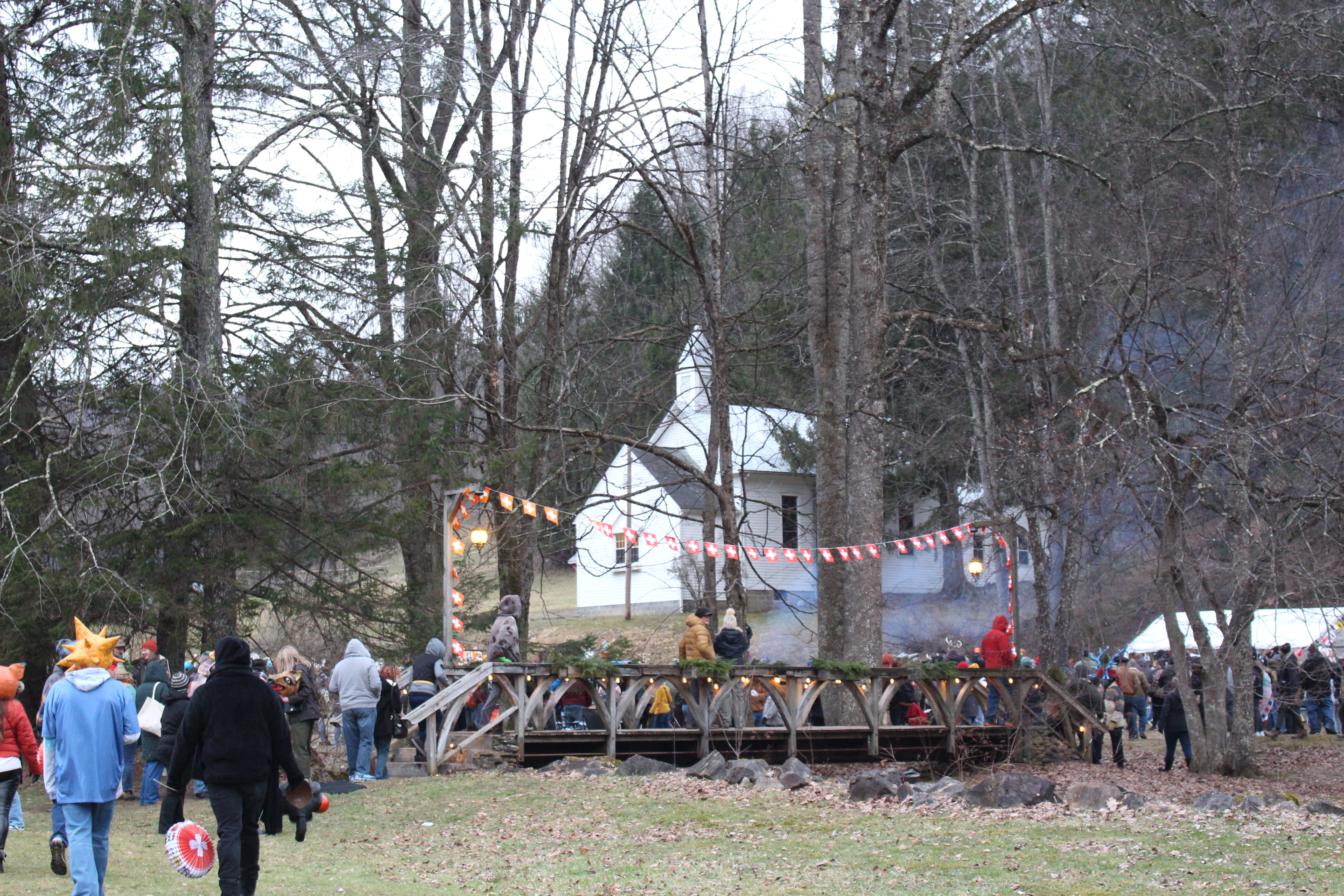 A white chapel in the background of a wooded place. In the foreground, people wearing handmade masks and winter coats walk together to cross a wooden bridge. A string of Swiss flags hang overhead the bridge.