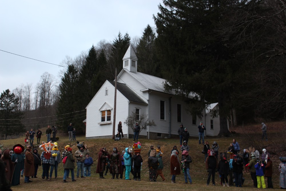 A white chapel in the background on an overcast evening. Several people stand in the foreground in a line wearing handmade masks.
