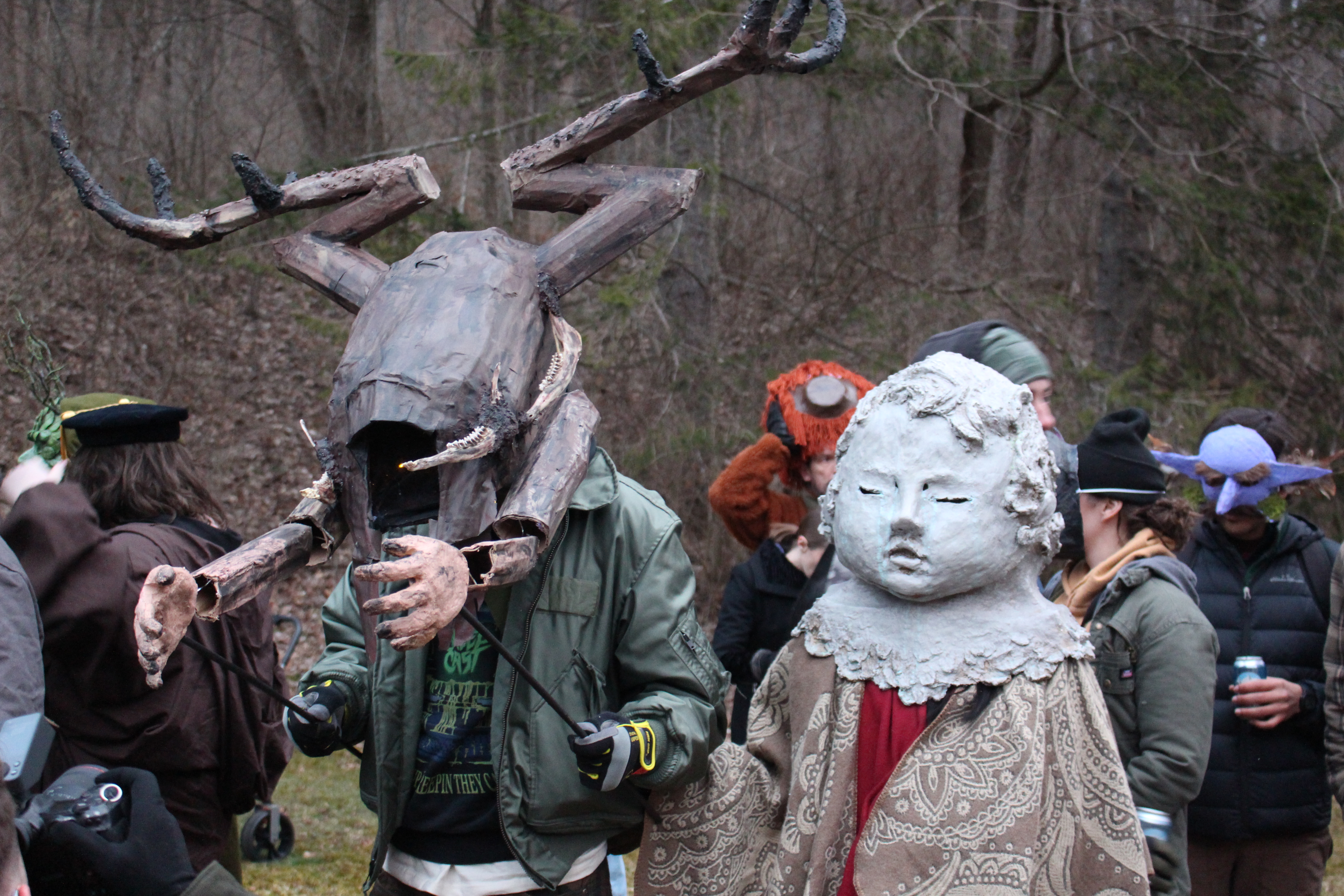 People in costumes gather outdoors during Fasnacht for the costume contest. This image is of two people. One wearing a distorted brown creature on their head with fake hands. The other looks to be a statue baby costume.