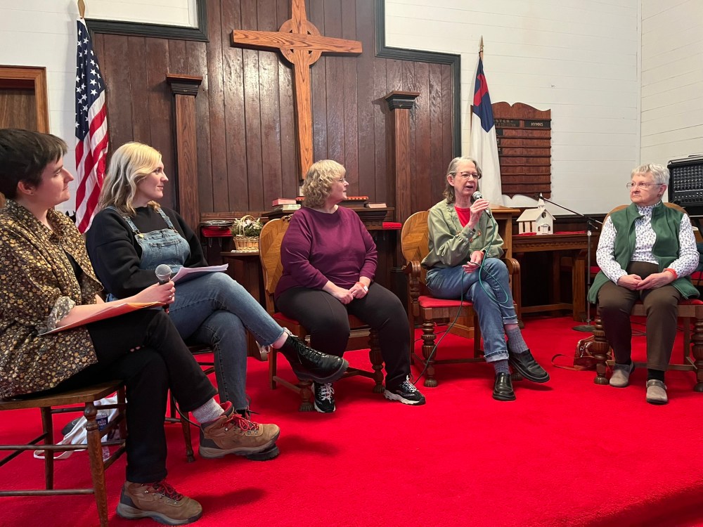 Five women sit in a semi-circle row at the alter of a small chapel. The carpet is bright red and there is a wooden cross behind them with two flags. Two younger women sit on the left two seats, one is holding a microphone. Three older women sit in chairs to the right. The one in the center is holding a microphone and speaking while the rest watch her.
